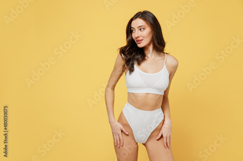 Smiling lovely attractive young brunette woman 20s in white underwear with perfect fit body standing posing look aside on workspaece area mock up isolated on plain yellow background studio portrait