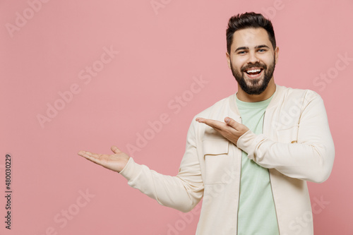 Young smiling happy european man 20s wearing trendy jacket shirt point hands arms aside on copy space area mock up workspace isolated on plain pastel light pink background. People lifestyle concept.