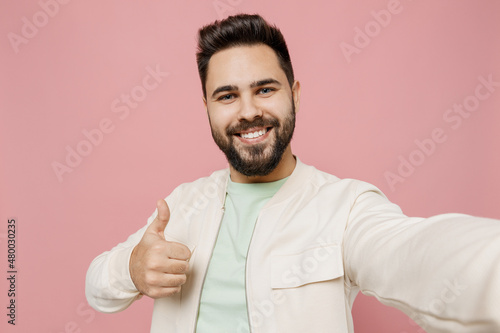 Close up young smiling happy man 20s in trendy jacket shirt doing selfie shot pov on mobile phone show thumb up gesture isolated on plain pastel light pink background studio People lifestyle concept