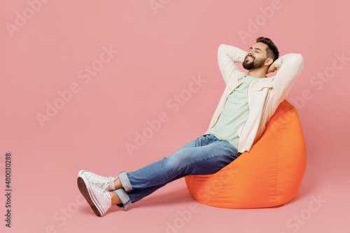 Full body young smiling happy man 20s wear trendy jacket shirt sit in bag chair hold hands behind neck sleep isolated on plain pastel light pink background studio portrait. People lifestyle concept