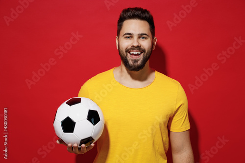Happy young bearded man football fan in yellow t-shirt cheer up support favorite team hold soccer ball isolated on plain dark red background studio portrait. People sport leisure lifestyle concept