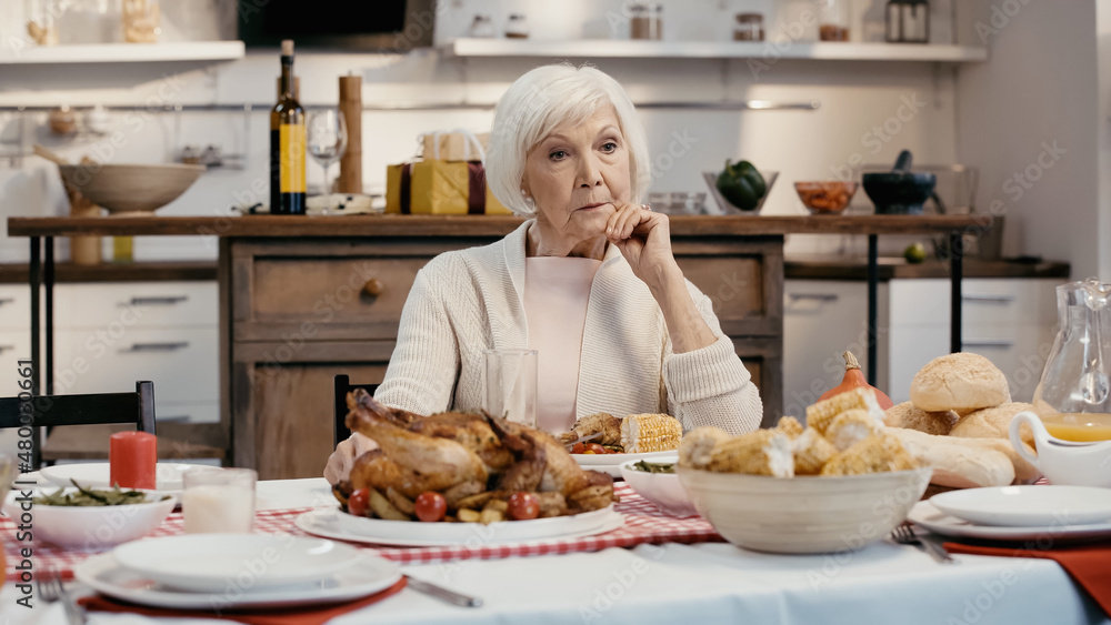 lonely and thoughtful senior woman sitting near thanksgiving dinner served on table in kitchen.