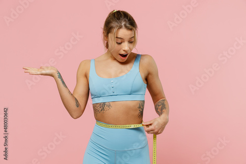 Young amazed strong sporty athletic fitness trainer instructor woman in blue tracksuit spend time in home gym hold measure tape on waist isolated on pastel plain pink background Workout sport concept.