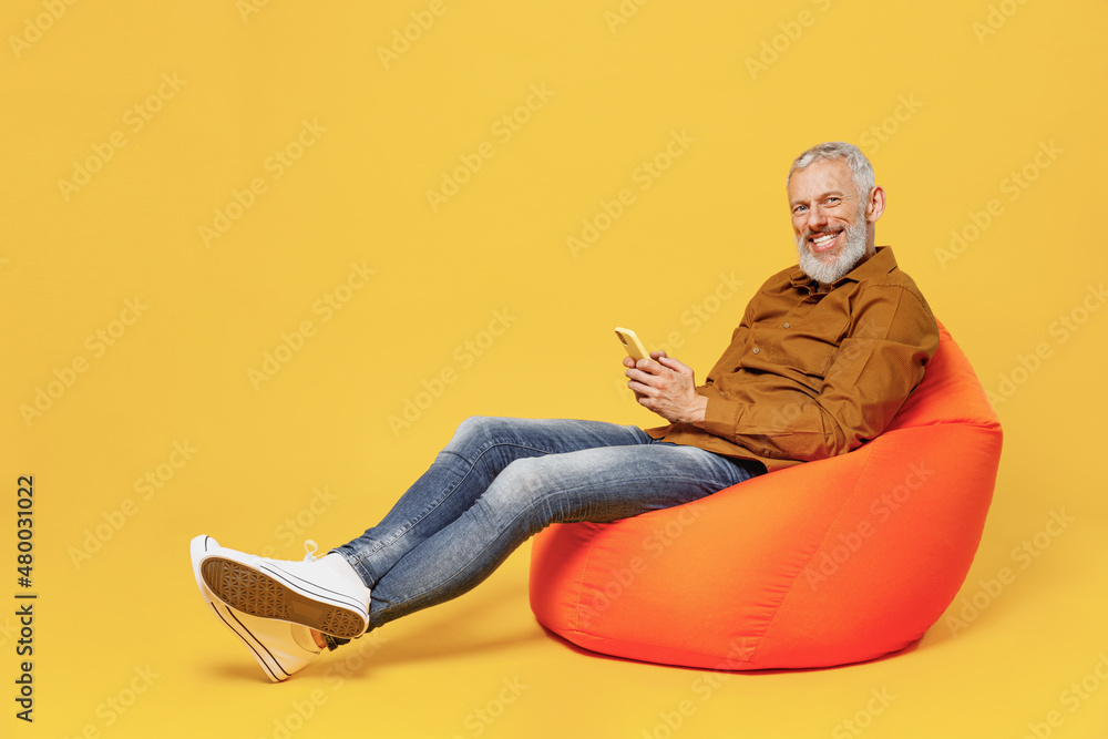 Full size body length smiling elderly gray-haired bearded man 40s years old wears brown shirt sit in bag chair hold in hand use mobile cell phone isolated on plain yellow background studio portrait. Full size body length smiling elderly gray-haired bearded man 40s years old wears brown shirt sit in bag chair hold in hand use mobile cell phone isolated on plain yellow background studio portrait.