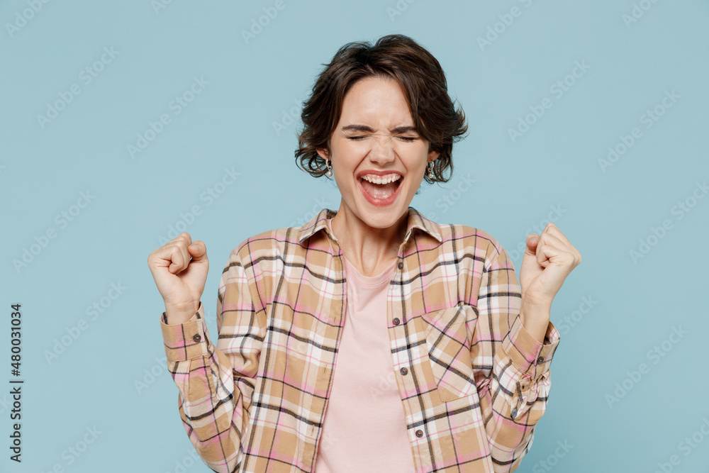 Young overjoyed caucasian woman 20s wear casual brown shirt doing winner gesture celebrate clenching fists say yes isolated on pastel plain light blue color background studio People lifestyle concept.