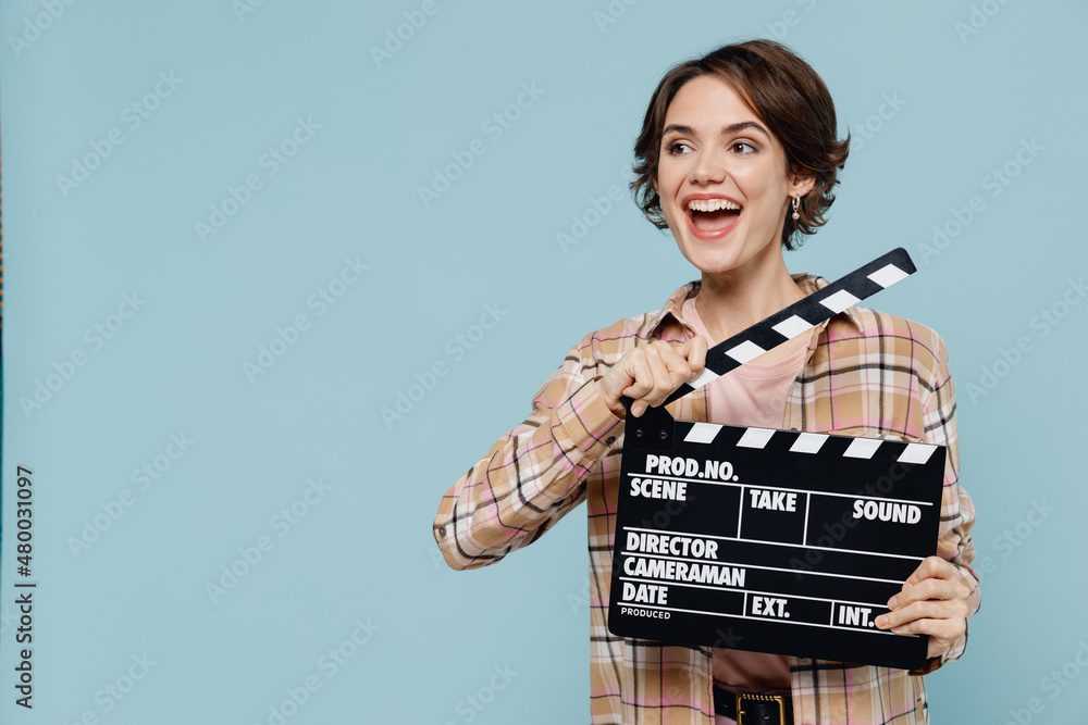 Young smiling happy woman 20s wearing casual brown shirt holding classic black film making clapperboard look aside on workspace isolated on pastel plain light blue color background studio portrait