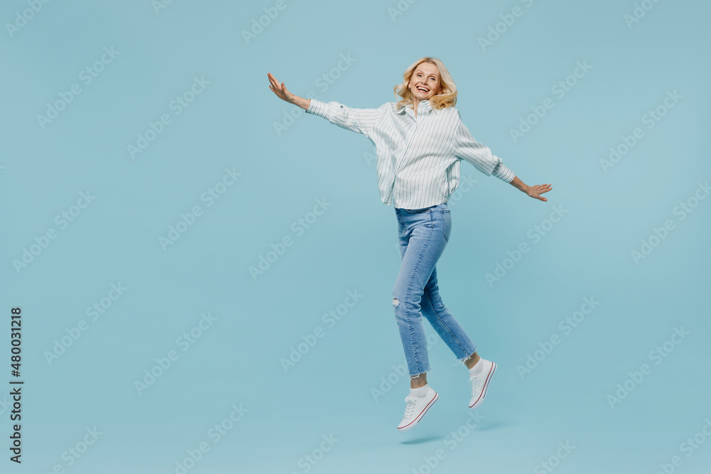 Full body elderly woman 50s wearing casual striped shirt looking camera jump hilg with outstretched hands like fly run isolated on plain pastel light blue color background. People lifestyle concept