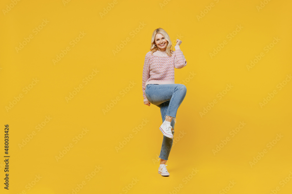 Full body elderly fun excited blonde overjoyed woman 50s in pink sweater do winner gesture clench fist raise up leg isolated on plain yellow color background studio portrait. People lifestyle concep. Full body elderly fun excited blonde overjoyed woman 50s in pink sweater do winner gesture clench fist raise up leg isolated on plain yellow color background studio portrait. People lifestyle concep.