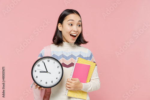 Smiling cool teen student girl of Asian ethnicity wear sweater backpack hold books clock look aside on workspace isolated on pastel plain light pink background Education in university college concept