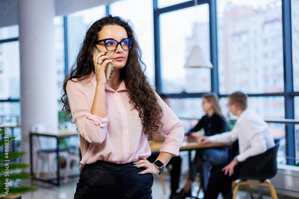 Smart happy young business woman, in stylish clothes and glasses, works in the office, talks with a client on a smartphone, gestures, smiles. Employees are working in the background. Selective focus