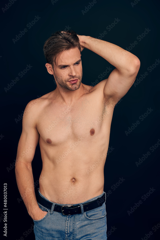 Handsome young bearded man isolated. Topless muscular man in jeans posing on dark blue background. Serious man looking at camera Handsome young bearded man isolated. Topless muscular man in jeans posing on dark blue background. Serious man looking at camera