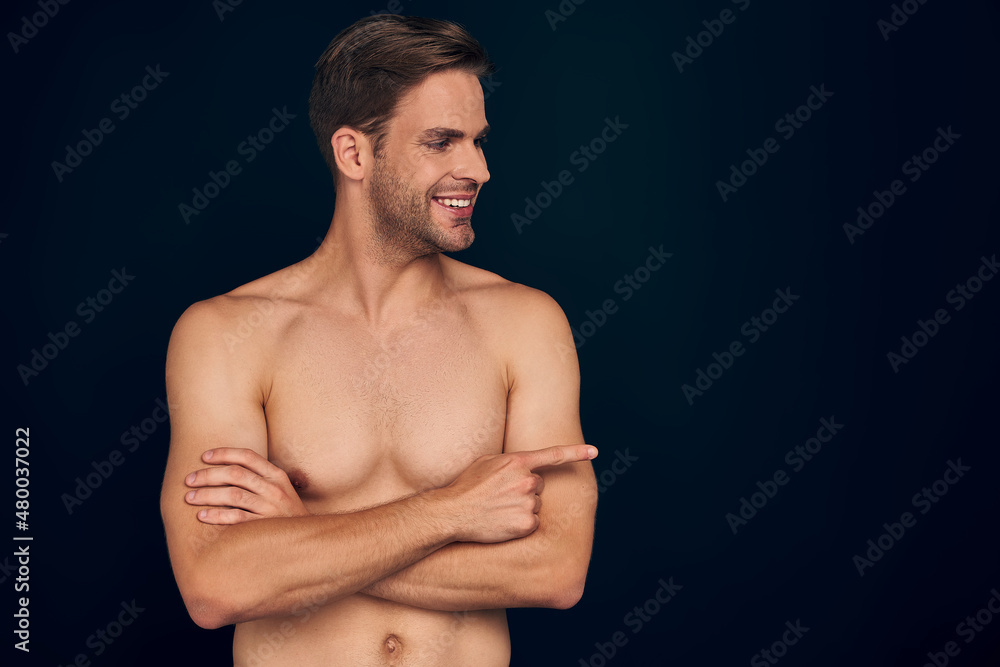 Fototapeta premium Handsome young man isolated. Portrait of shirtless muscular man is standing on dark blue background and pointing to the side. Showing product.