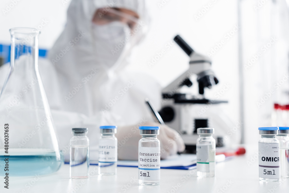 Fototapeta premium selective focus of vials with coronavirus vaccine near scientist writing near microscope on blurred background.