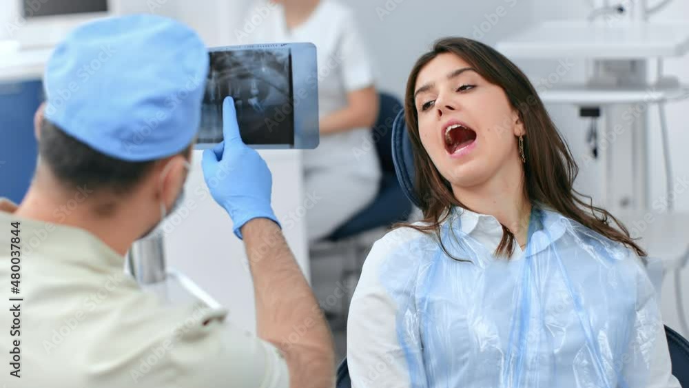 Male dentist pointing on x ray image consulting female patient open mouth on chair at stomatology clinic Male dentist pointing on x ray image consulting female patient open mouth on chair at stomatology clinic