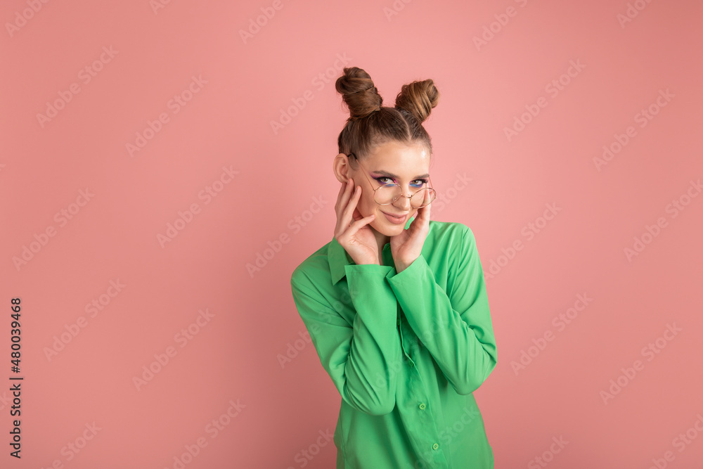 Young pretty stylish woman in green shirt and round glasses with two bun hairstyles on pink background. Beauty and fashion. Copy space Young pretty stylish woman in green shirt and round glasses with two bun hairstyles on pink background. Beauty and fashion. Copy space