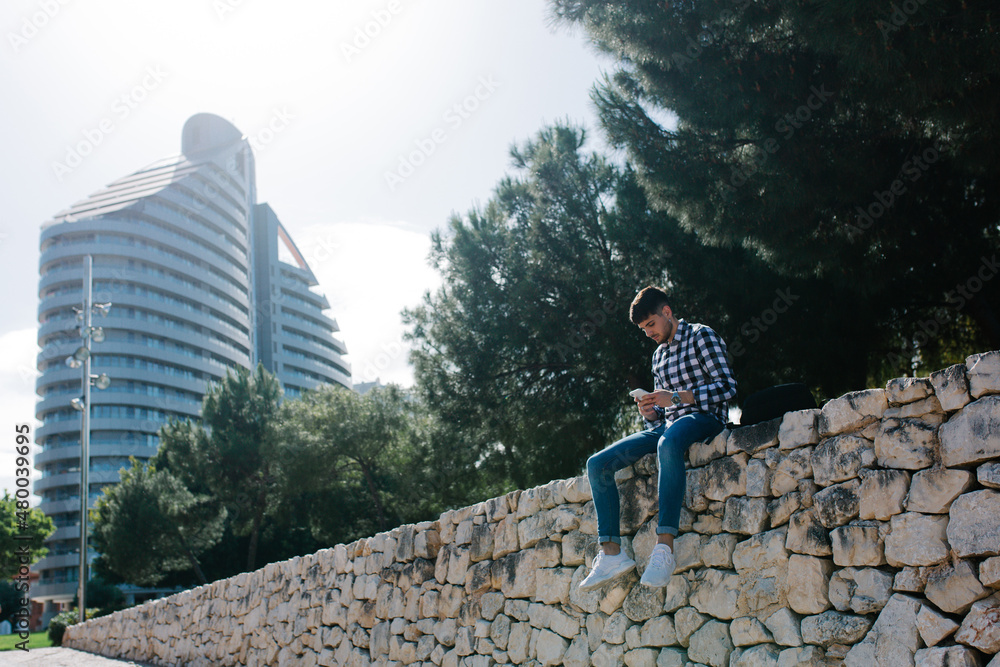 Young man sitting on a stone wall using his cell phone