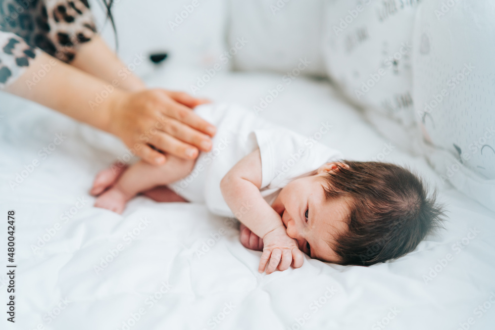 Cute little newborn baby with dark hair in white bodysuit lie sidelong smiling and mother touching his legs. Copy space. Cute little newborn baby with dark hair in white bodysuit lie sidelong smiling and mother touching his legs. Copy space.