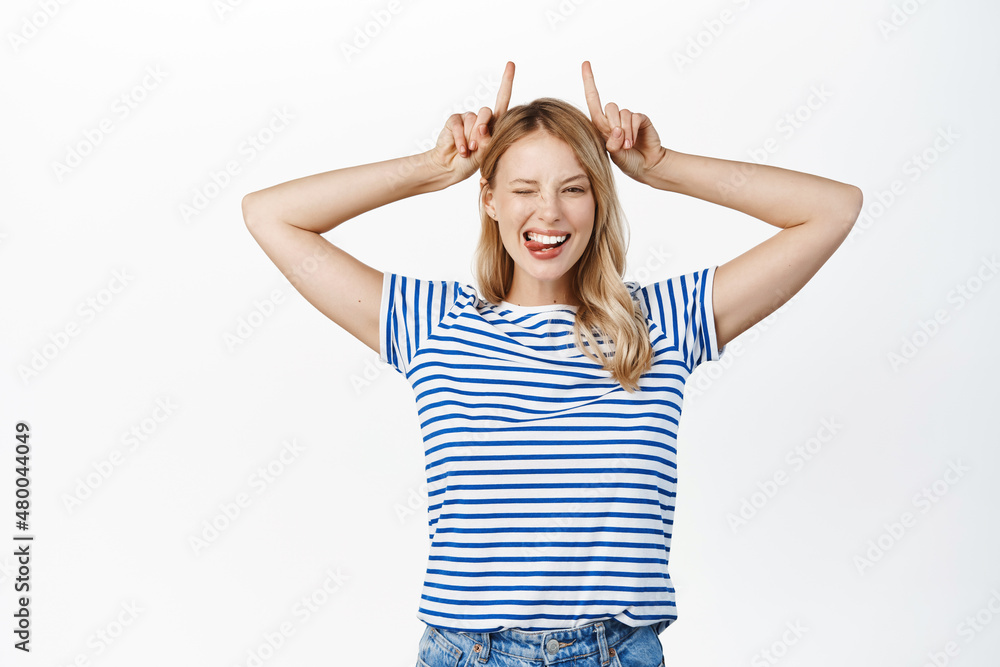 Happy stylish girl shows bull horns, pointing fingers up on her head and smiling, winking at camera, showing promo advertisement, standing over white background