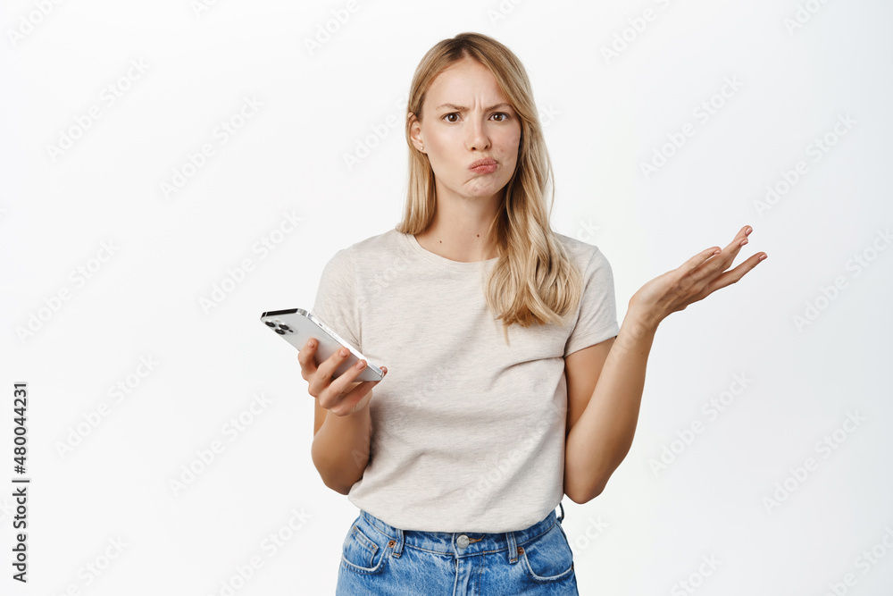 Confused blond woman shrugging with mobile phone, cant understand smth on smartphone, standing puzzled over white background Confused blond woman shrugging with mobile phone, cant understand smth on smartphone, standing puzzled over white background