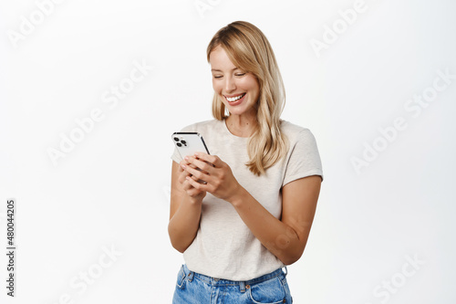 Cellular technology, mobile apps concept. Smiling beautiful woman using smartphone, looking at her phone with happy face, reading, standing over white background