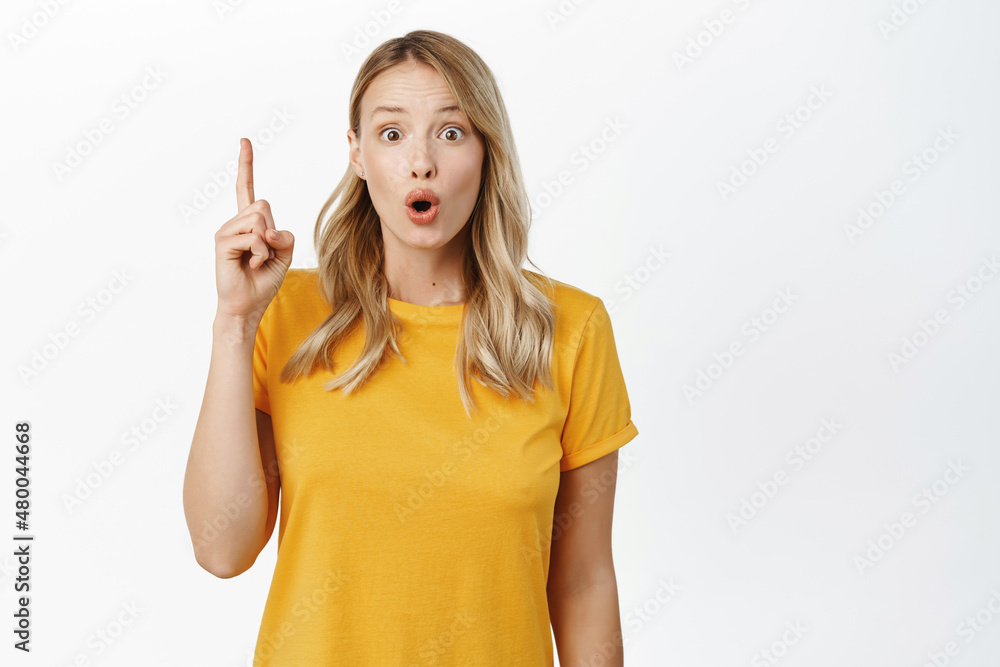 Amazed blond girl showing advertisement, pointing finger up, demonstrating something on top, logo or banner ahead, white background