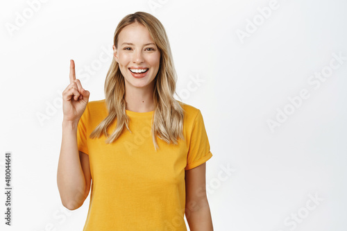 Smiling young blond girl showing advertisement  pointing finger up  demonstrating promo offer  recommending company  standing over white background