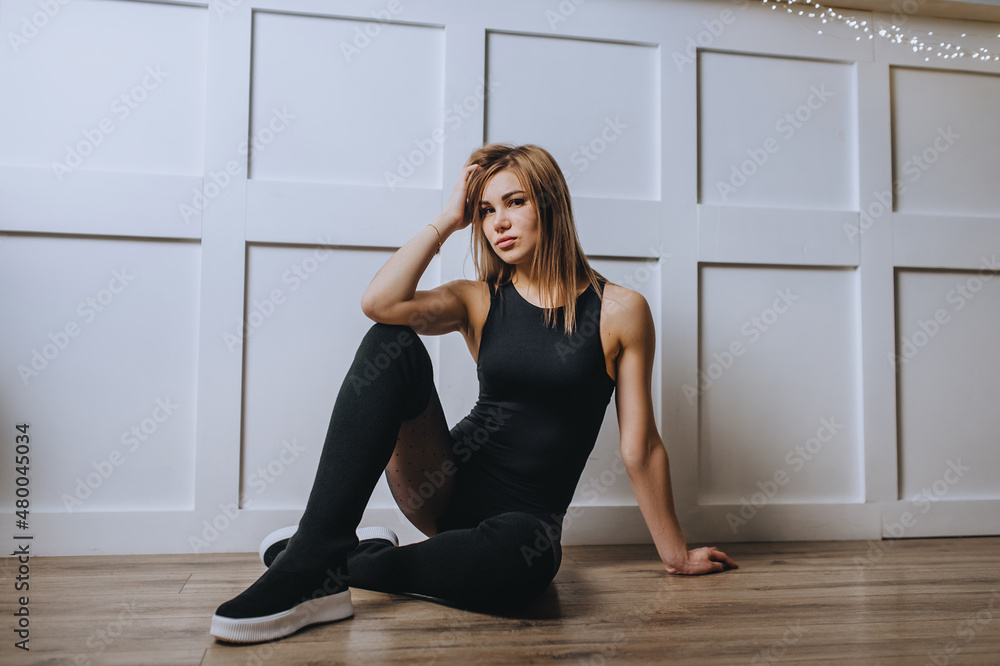 Beautiful girl athlete blonde in black sportswear sits near a white wall in the interior.