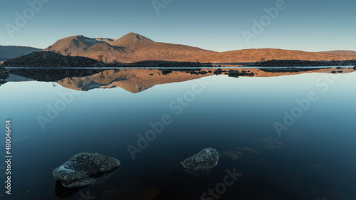 The tops of the mountains reflected in the surface of the lake with two stones in the background.