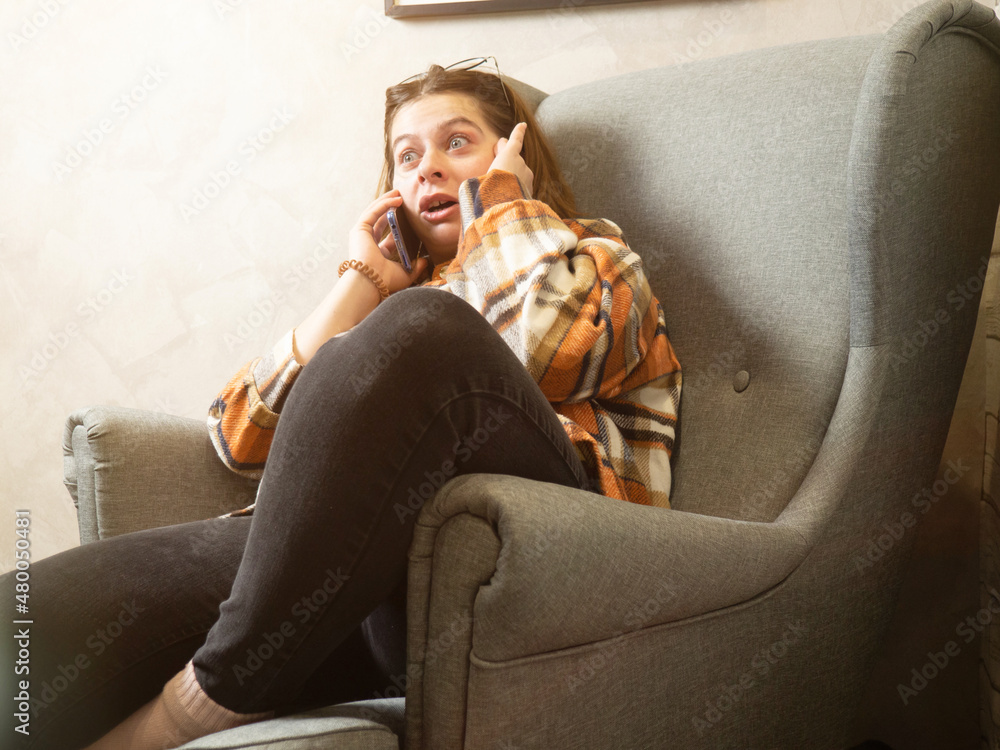 Obraz premium a girl is talking on a mobile phone at home in an armchair, her eyes bulging out that something proves to the interlocutor