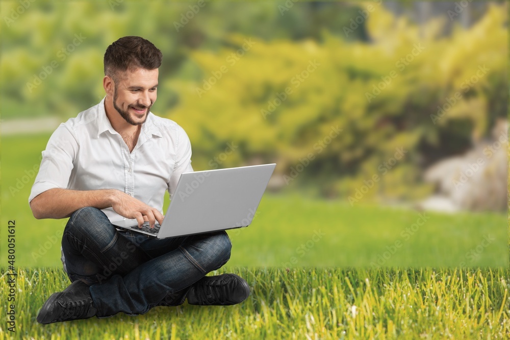 Young businessman student freelancer using laptop for doing homework, on green grass lawn Young businessman student freelancer using laptop for doing homework, on green grass lawn
