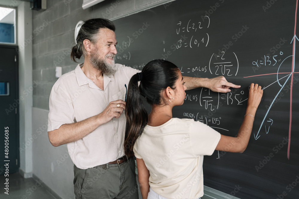 Maths lesson in a secondary school. Latin Female Teenager Student writing on Blackboard with help of friendly teacher in High School. Maths lesson in a secondary school. Latin Female Teenager Student writing on Blackboard with help of friendly teacher in High School.