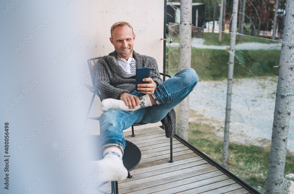 Smiling middle-aged man dressed open cardigan, jeans, and warm socks sitting on forest house balcony and using modern smartphone. Everyday lifestyle photo with modern devices concept image..