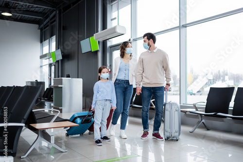 family in medical masks walking with baggage in airport lounge.
