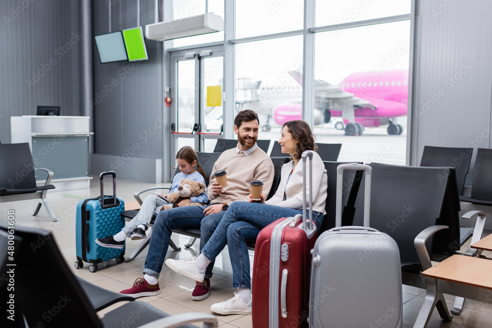happy parents with paper cups near daughter and luggage in airport lounge. happy parents with paper cups near daughter and luggage in airport lounge.
