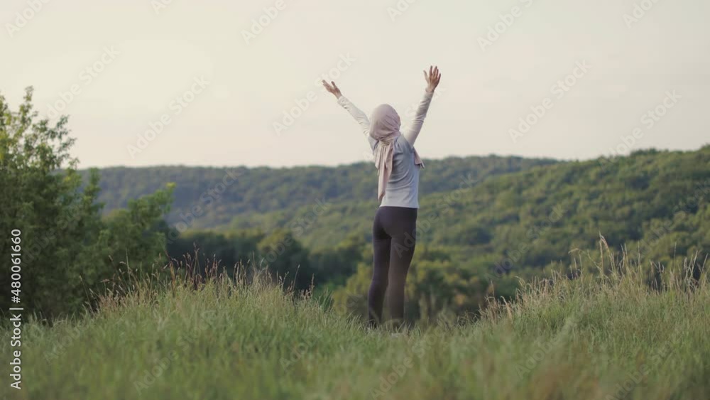 An Arabian young woman is practicing yoga in the summer park. A Muslim woman is looking aside exercising outdoor and breathing fresh air outstretching arms . Fit arabic woman training in a park An Arabian young woman is practicing yoga in the summer park. A Muslim woman is looking aside exercising outdoor and breathing fresh air outstretching arms . Fit arabic woman training in a park