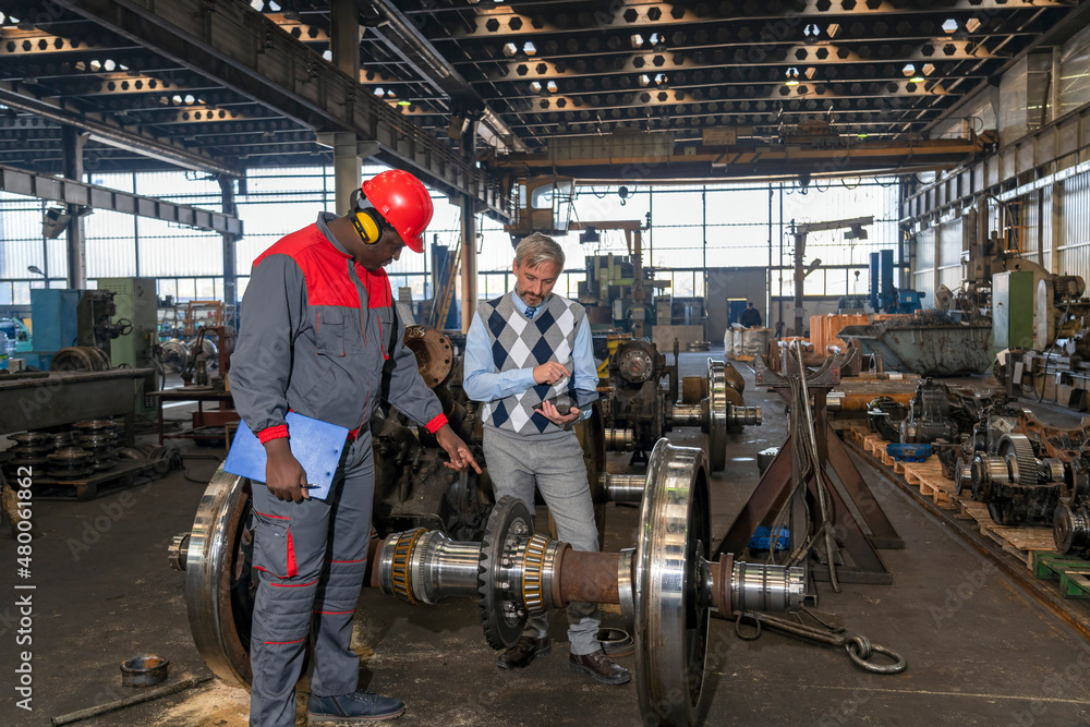 Production Supervisor Talking To African American Worker Next To Train Wheels At Train Factory. Area For Maintenance, Repair And Service Of Trains And Wagons. Production Supervisor Talking To African American Worker Next To Train Wheels At Train Factory. Area For Maintenance, Repair And Service Of Trains And Wagons.