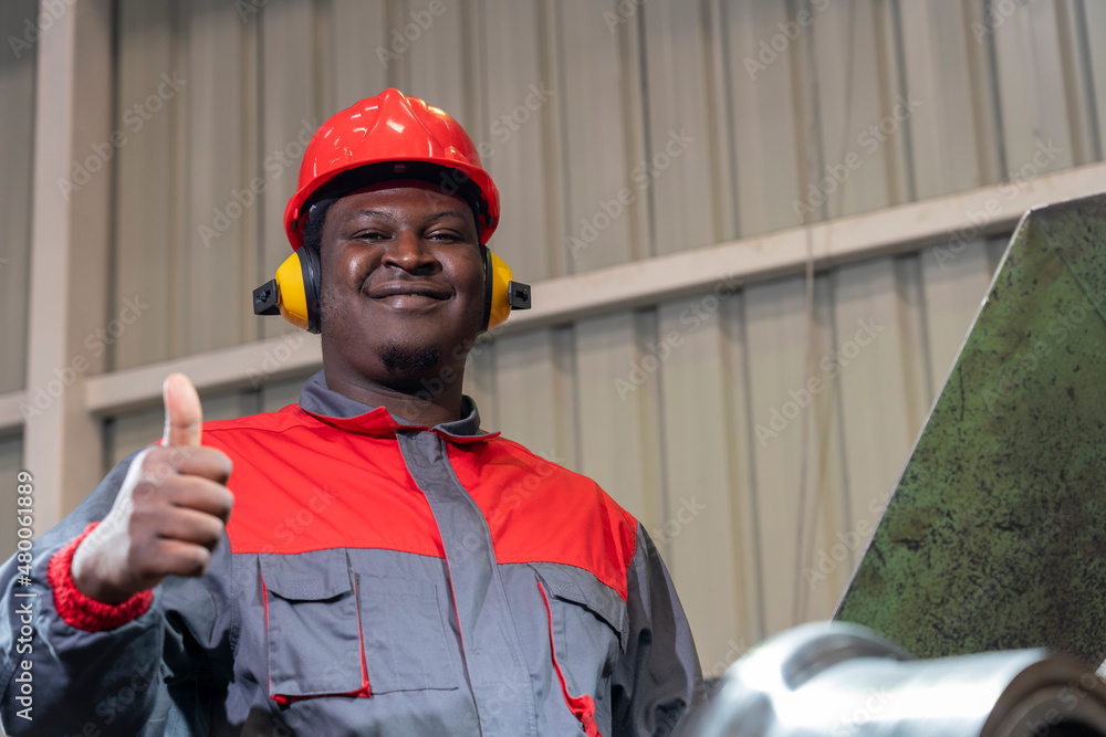 Smiling African American Blue-Collar Worker Giving Thumbs Up In A Factory.  Portrait Of Black Worker In Red Helmet, Hearing Protectors And Work Uniform  . Photos | Adobe Stock