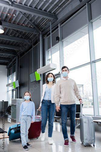 family in medical masks walking with suitcases in airport lounge.