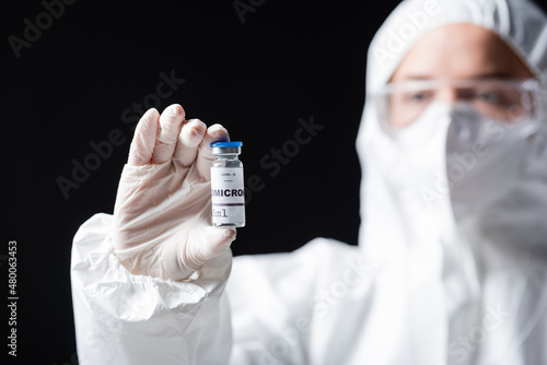 blurred immunologist in white hazmat suit holding covid-19 omicron variant vaccine isolated on black.
