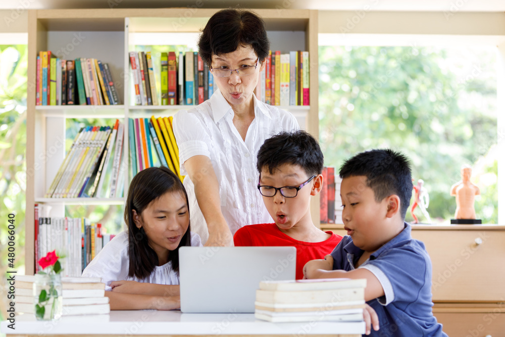 A Group of Asian Student Kid Reading and looking Laptop on the table with female teacher in School library with Shelf of Books Background A Group of Asian Student Kid Reading and looking Laptop on the table with female teacher in School library with Shelf of Books Background