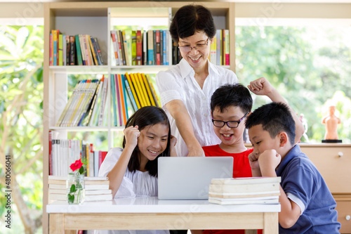A Group of Asian Student Kid Reading and looking Laptop on the table with female teacher in School library with Shelf of Books  Background