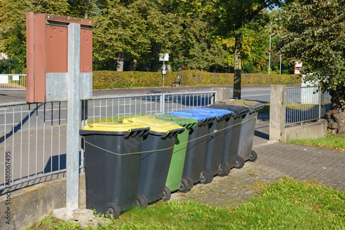 A group of plastic trash and recycle bins by a metal fence. © Viktor