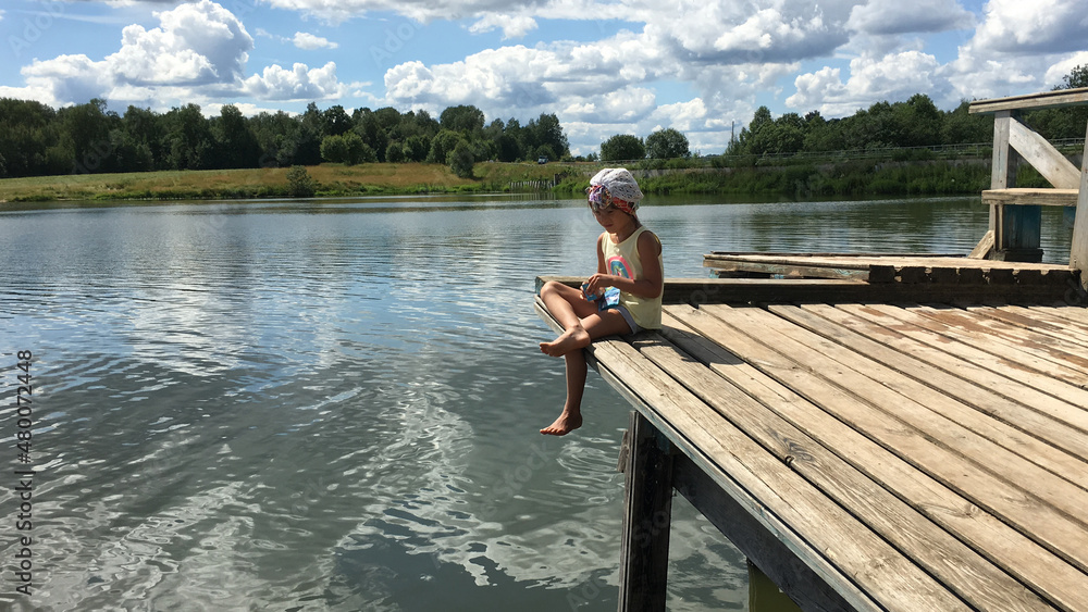 funny country alone barefoot child in cap sits on the edge of a wooden bridge over the water and dream. natural background sky with clouds funny country alone barefoot child in cap sits on the edge of a wooden bridge over the water and dream. natural background sky with clouds