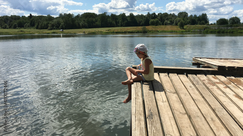 funny country alone barefoot child in cap sits on the edge of a wooden bridge over the water and dream. natural background sky with clouds