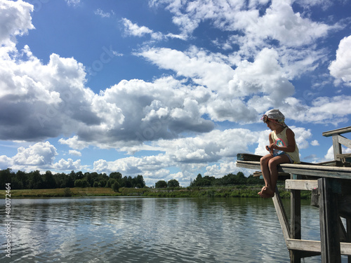 country alone barefoot child in cap sits on the edge of a wooden bridge over the water and watch. natural background sky with clouds © Natalya