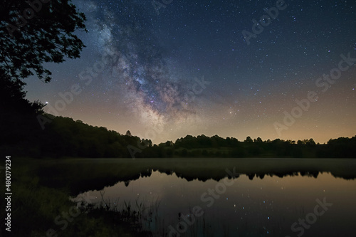 The Milky Way over volcanic lake of Weinfelder Maar in the Eifel with reflection of stars on water surface, Daun, Germany
