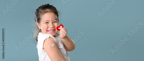 Coronavirus Vaccination Advertisement. Happy Vaccinated Little asian child girl Showing Arm With Plaster Bandage After Covid-19 Vaccine Injection Posing Over Blue Background, Smiling To Camera. 