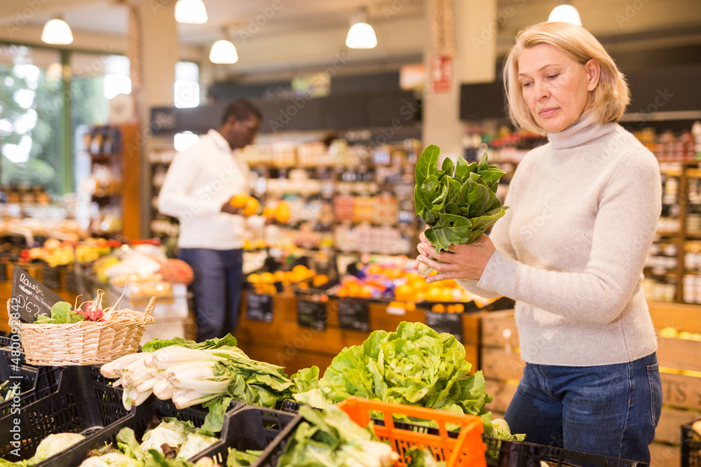 Focused middle aged woman shopping in organic food store, choosing fresh vegetables