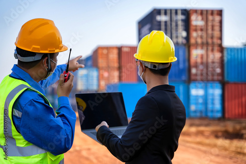 An Asian businessman in a black suit holds a laptop to inspect a container yard.