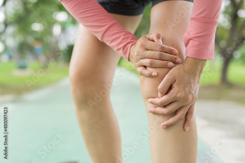 A young woman in sports outfits pink injured her knee during exercise in the park. Low section of sports girl suffering from joint pain while standing on track during. Accident from exercise concept.